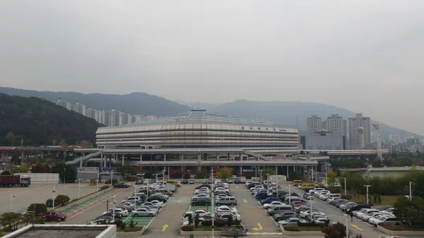 Archival exterior view of Sajik Indoor Gymnasium in Busan, with mountains and a parking lot in the foreground.