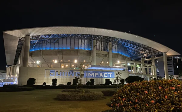 Nighttime exterior of IMPACT Arena in Muang Thong Thani, with its curved roof and blue-lit entrance against a dark sky.